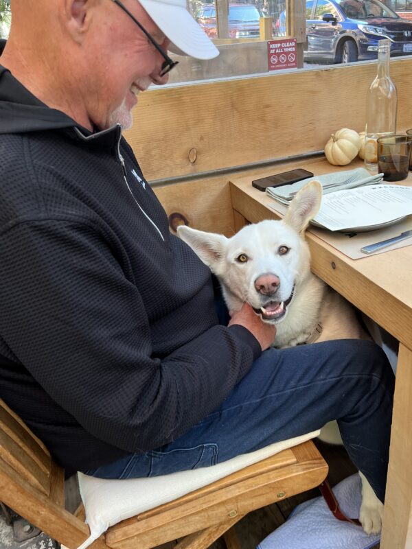 Man Sitting At Outdoor Table With Cute White Mutt Between Him And The Table